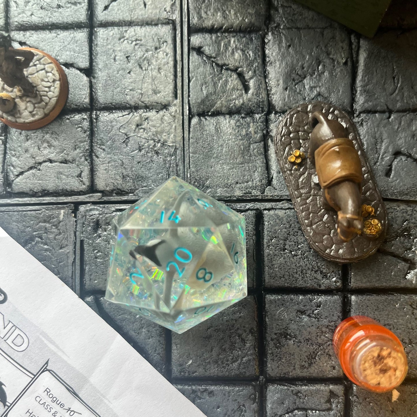 Clear resin D20 chonk showing the 20 face, with a toy penguin inside. The chonk has iridescent mylar tubes and glitter at the bottom, and is inked in ice blue. Shown on a gaming background.