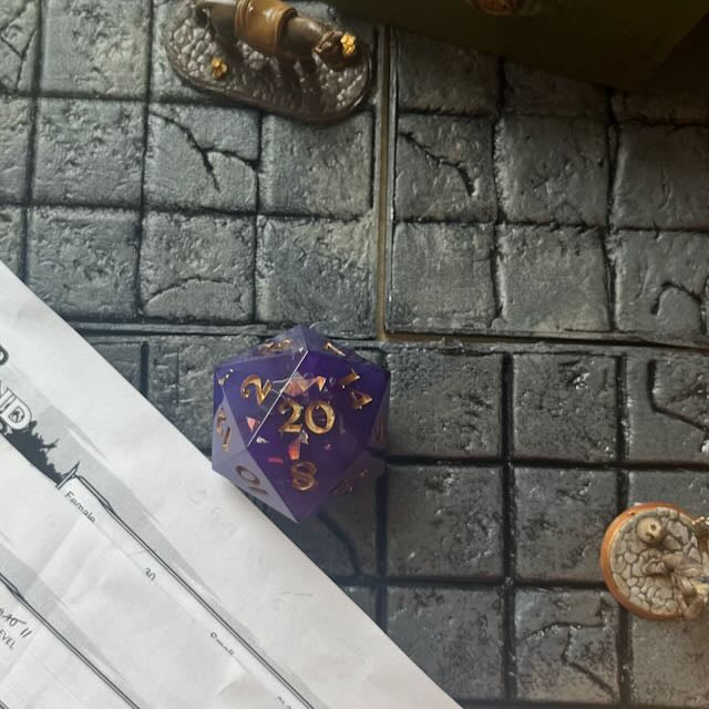 Opaque purple D20 chonk with glittery shiny mylar flakes, inked in gold on a D&D table