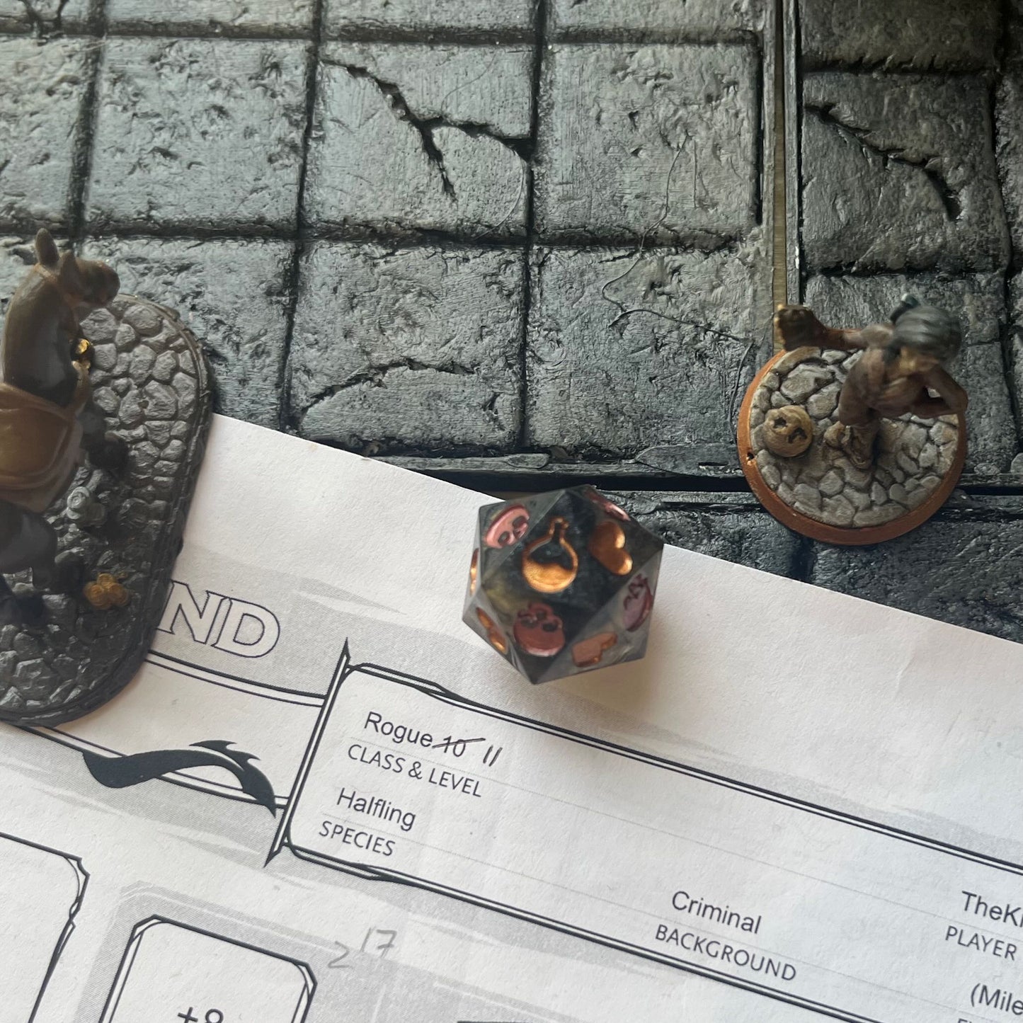 Resin D20 dice used for Death Saves in TTRPG. Semi transparent gold and black, skulls inked in copper and hearts inked in gold shown on a D&D background
