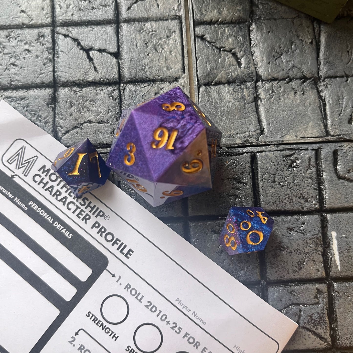 Full set of Mothership dice on a Mothership background. The D20 is a chonk, and the D10 and D100 are regular size. The resin is coloured with pigment powders using black, blue and red. The dice are inked in gold.