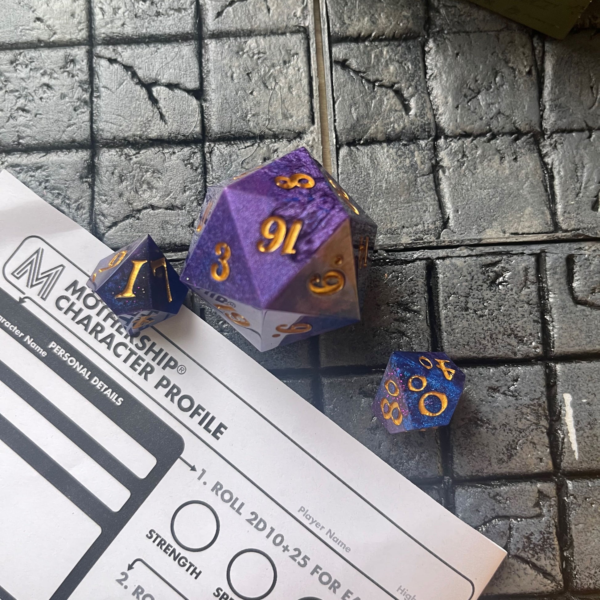 Full set of Mothership dice on a Mothership background. The D20 is a chonk, and the D10 and D100 are regular size. The resin is coloured with pigment powders using black, blue and red. The dice are inked in gold.