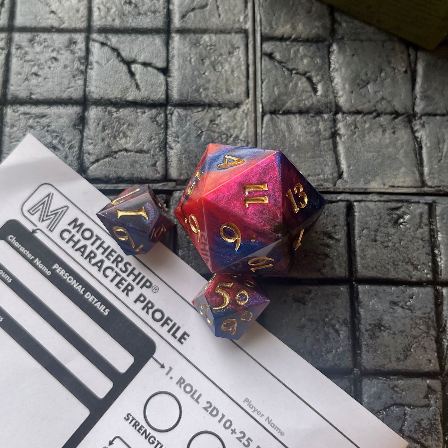 Full set of Mothership dice shown on a gaming background. The D20 is a chonk, and the D10 and D100 are regular size. The resin is coloured with pigment powders using black, blue and red. The dice are inked in chromed gold.