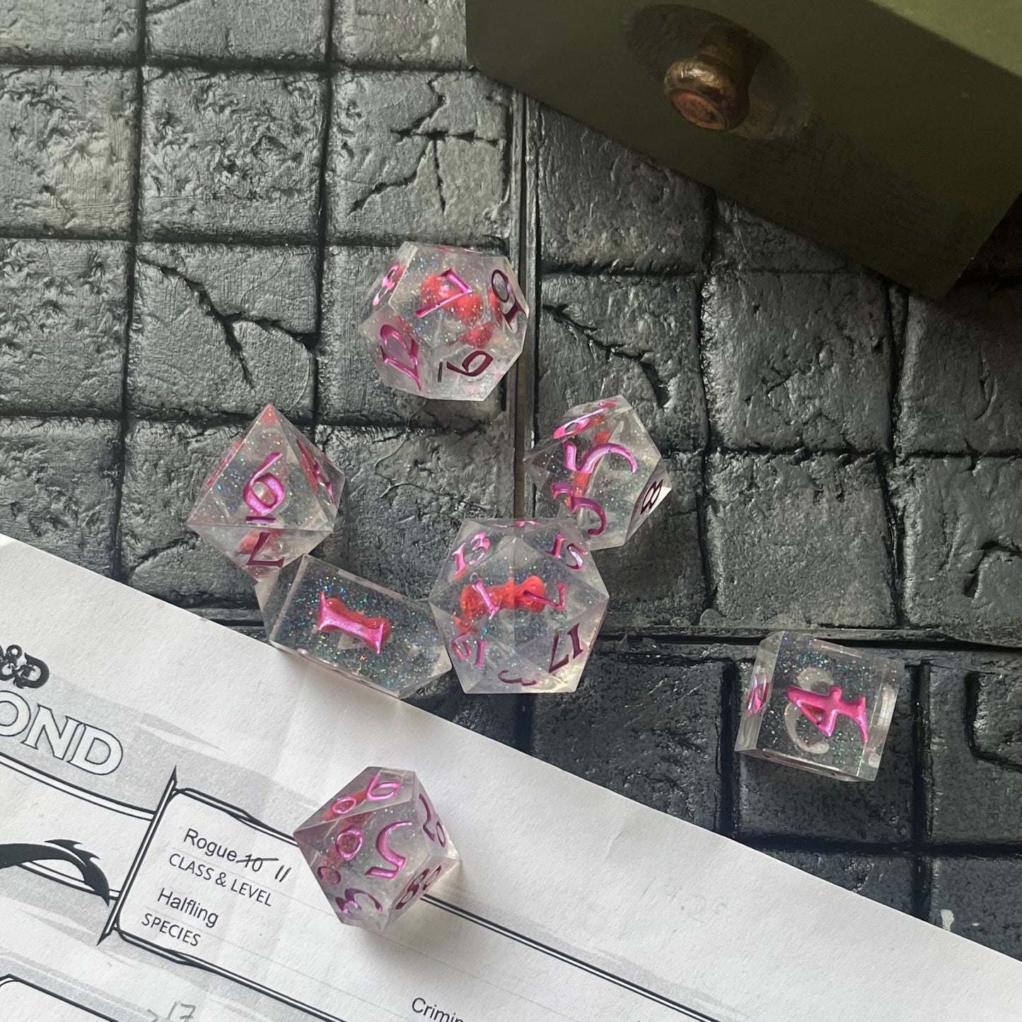 Full set of TTRPG resin dice shown on a D&D background. Resin is transparent with translucent glitter suspended inside and a red 3D printed hair bow. These are inked in colour-shifting pink pigment