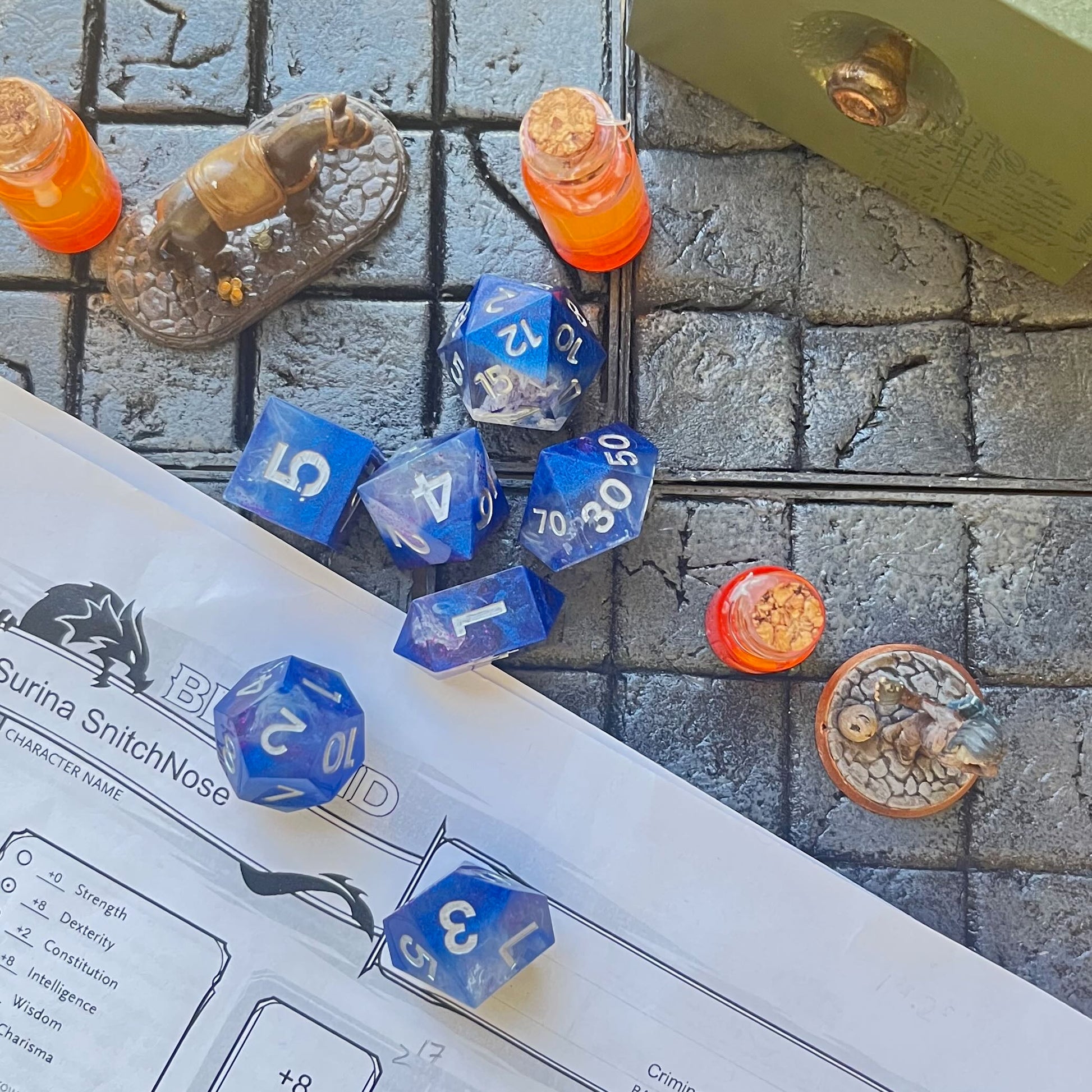 Showing Full set of TTRPG resin dice, opaque blue in parts, with clear areas with white resin. Inked in silver. On a D&D background