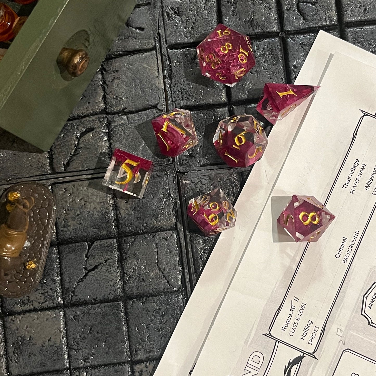 Full set of TTRPG resin dice, petri-pour in white and Sweet Cherry, inked in Gold on a D&D background