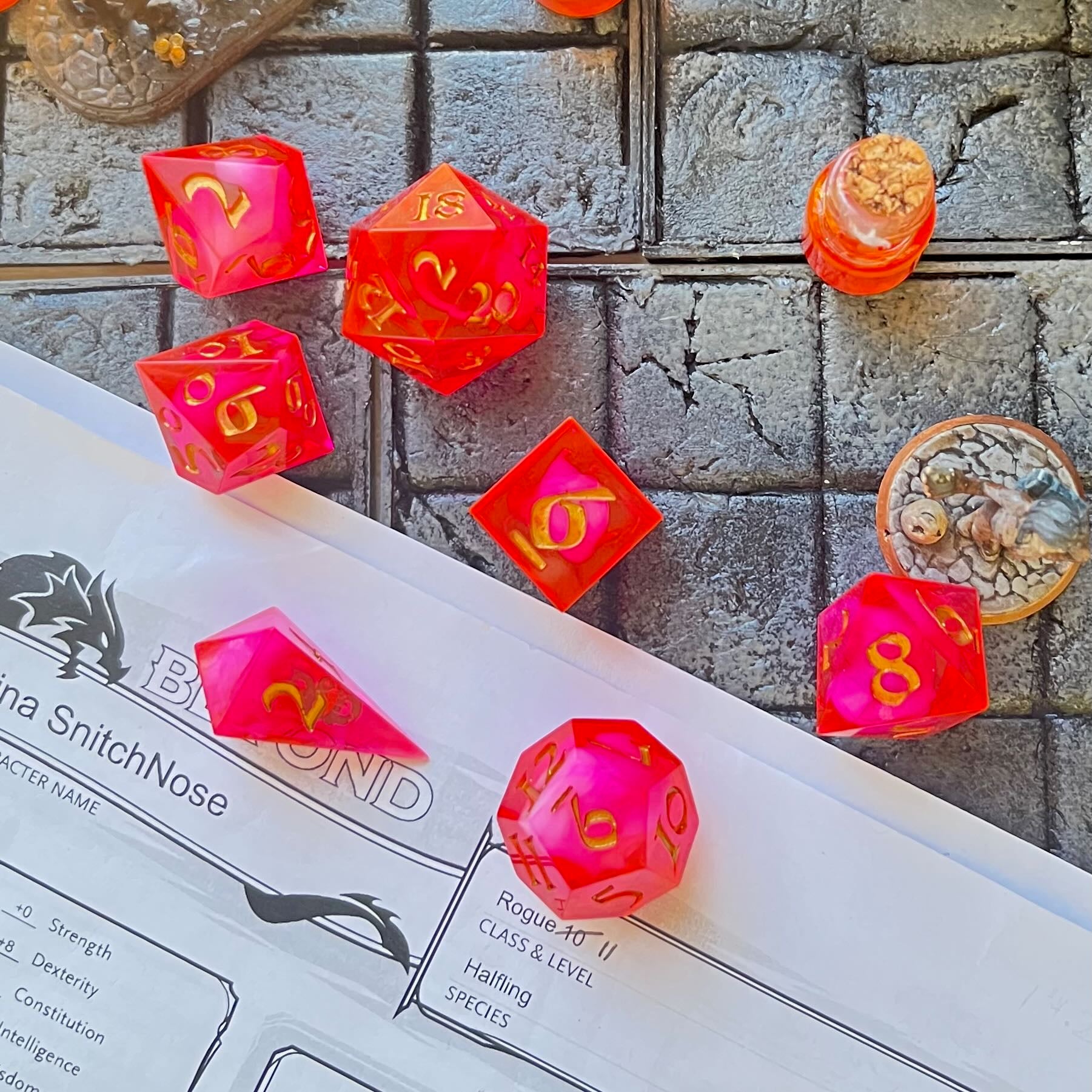 Full set of TTRPG semi-transparent resin dice, coloured with red and pink dye, with a soft blur of resin coloured with Piñata Blanco Blanco white in the centre and inked in gold. Shown on a D&D background