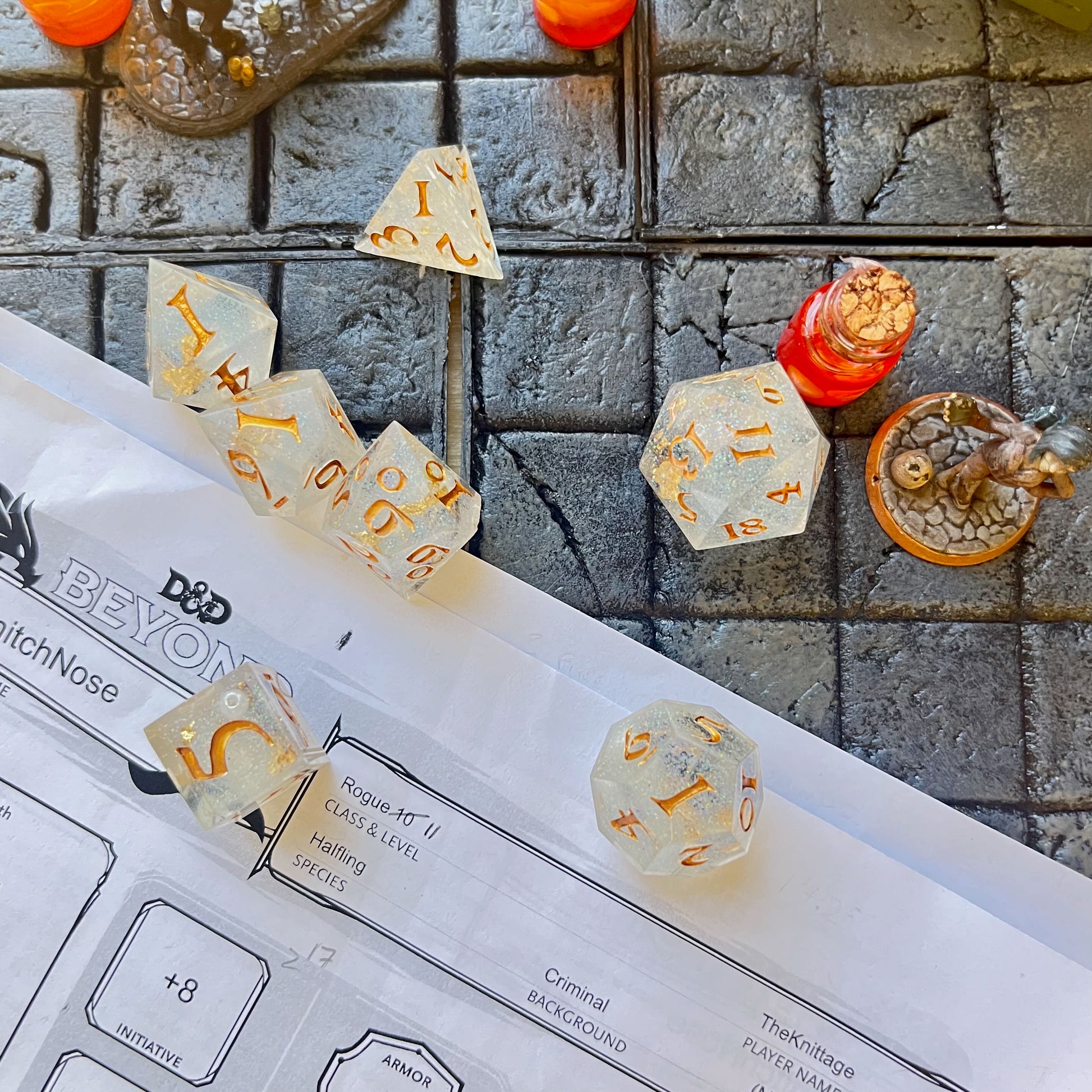 Full set of TTRPG dice, semi-transparent resin with translucent glitter and angel wing and pearl beads suspended inside. Inked in gold. Shown on a D&D background.
