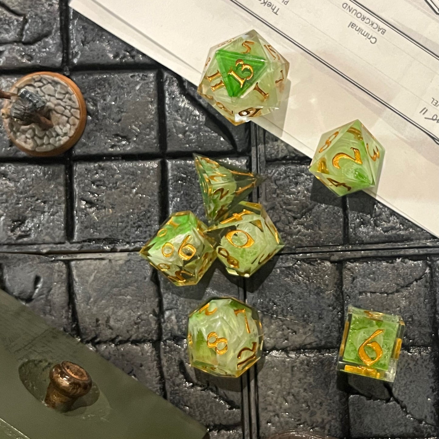 Full set of TTRPG resin dice, with green and white pigment cast into blanks and then shelled in clear resin and inked in Chromed Gold on a D&D background