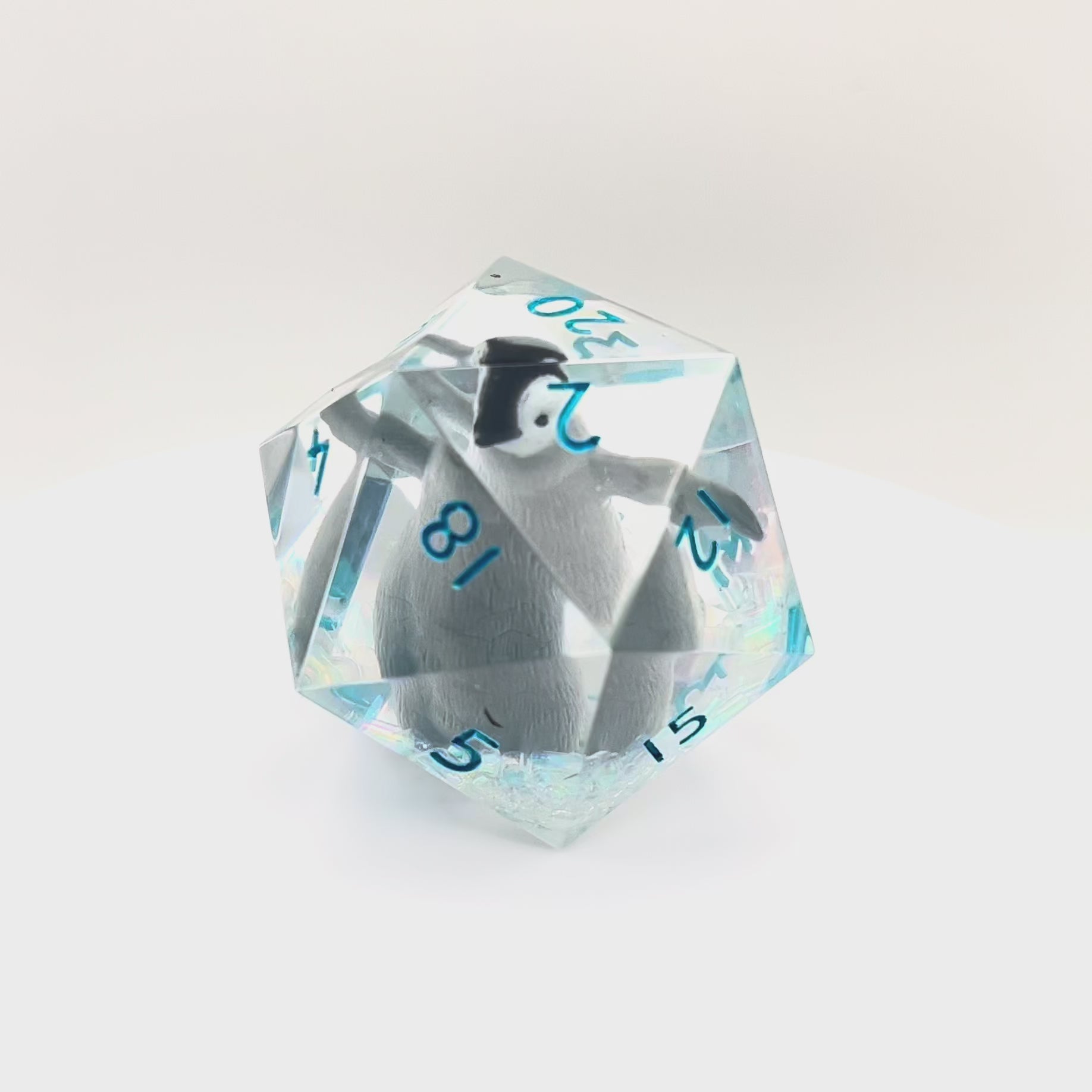 Video showing a clear resin D20 chonk with a toy penguin inside. The chonk has iridescent mylar tubes and glitter at the bottom, and is inked in ice blue