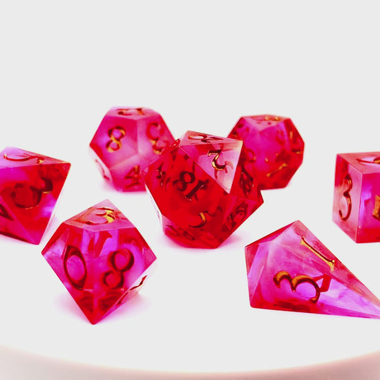 Video showing Full set of TTRPG semi-transparent resin dice, coloured with red and pink dye, with a soft blur of resin coloured with Piñata Blanco Blanco white in the centre and inked in gold