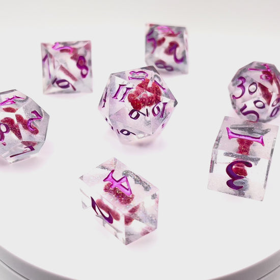 Video showing a full set of TTRPG resin dice. Resin is transparent with translucent glitter suspended inside and a red 3D printed hair bow. These are inked in colour-shifting pink pigment