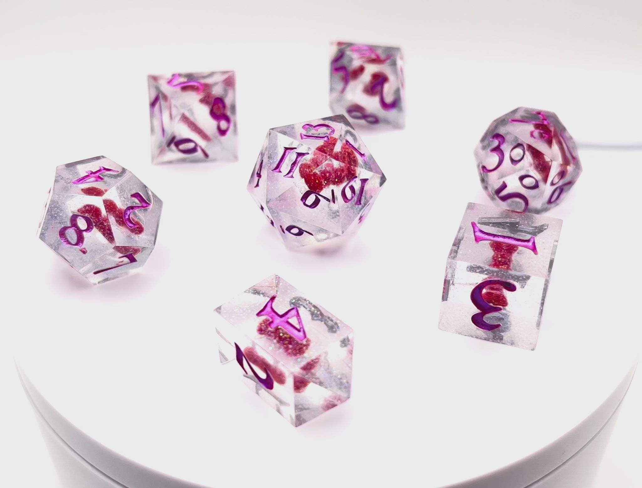 Video showing a full set of TTRPG resin dice. Resin is transparent with translucent glitter suspended inside and a red 3D printed hair bow. These are inked in colour-shifting pink pigment